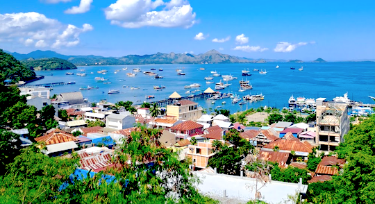 Located on the western tip of Flores Island, Indonesia, Labuan Bajo is a vibrant city, renowned as the gateway to Komodo National Park. This stunning aerial view captures the lively harbor area, filled with boats and liveaboards, set against a backdrop of rolling hills. As the sun sets, a brilliant golden glow spreads across the water, highlighting the city's bustling waterfront. Known for its tourism, adventure opportunities, and natural beauty, Labuan Bajo’s scenic landscapes attract visitors from around the world.